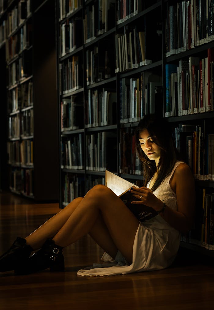 A woman reads a glowing book on the floor of a dimly lit library, creating a magical atmosphere.